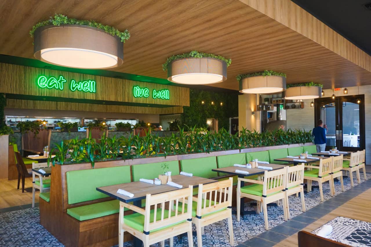 Food and Thought Cafe booths lined with plants above the seats and chairs along the aisle and neon signs "eat well" and "live well"